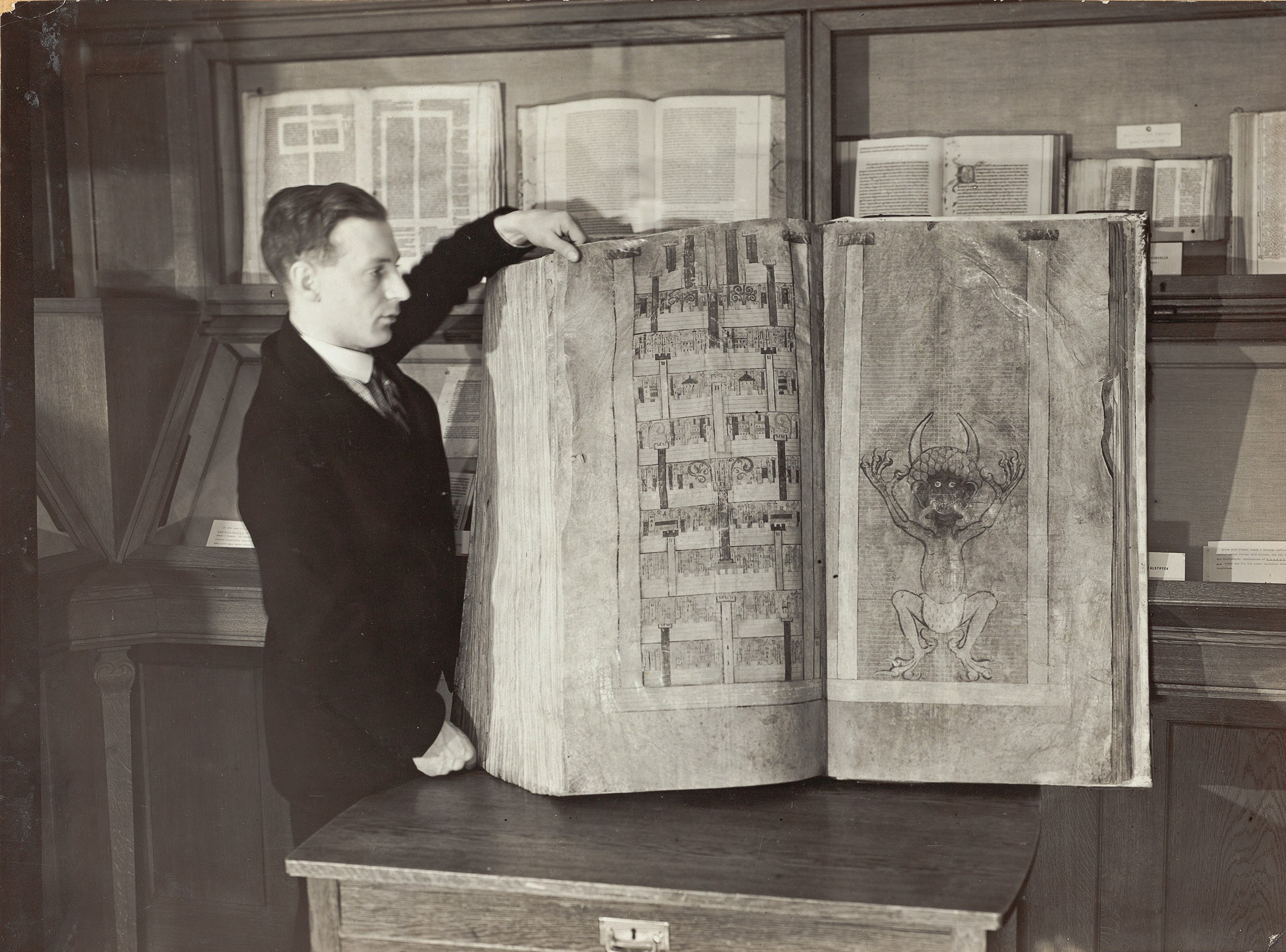 Svartvitt fotografi av en ung man iförd kostym som håller upp en stor bok med en bild av djävulen på ett bord framför en rad glasmontrar med böcker.
