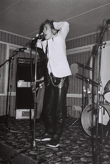 Svartvitt fotografi av en man i vit skjorta och skinnbyxor. Han håller händerna över öronen och sjunger i en mikrofon.